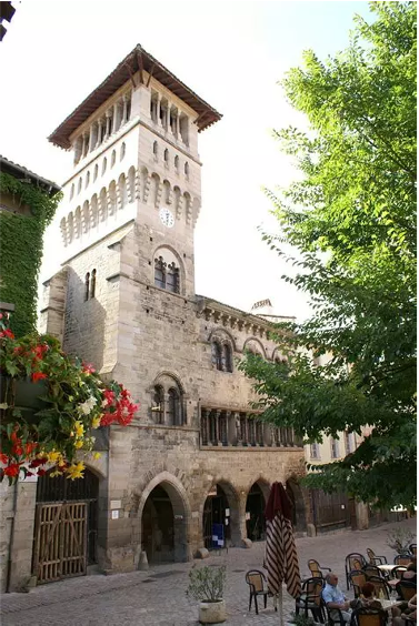 L'Îlot Muratet et la Maison Romane au centre de la vie de village à Saint-Antonin-Noble-Val. Façade complète de la Maison Romane et de la tour avec terrasse de café et végétation en avant-plan.